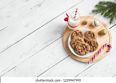 Chocolate Chip Cookies And Milk For Santa. Traditional Christmas Homemade Cookies, Candy Cane And Bottle Of Milk With Christmas Decor On White Wooden Table, Top View, Copy Space.