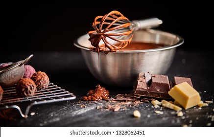 Chocolate coated whisk resting on mixing bowl filled with melted chocolate or sauce with a wire rack of bonbons and ingredients - Shutterstock ID 1328062451