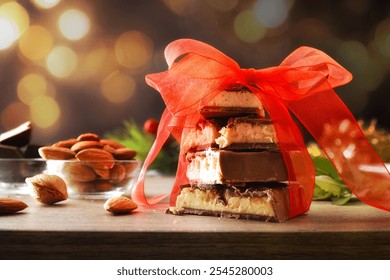 Chocolate covered nougat with stacked pieces tied with red ribbon on a wooden table with natural almonds and portions of chocolates and festive decoration and background with golden bokeh. - Powered by Shutterstock