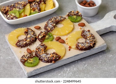Chocolate covered pineapple rings and kiwifruit slices sprinkled with chopped nuts and coconut flakes on white wooden board. Homemade healthy fruit treat. Soft focus, horizontal.