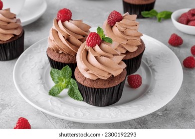 Chocolate cupcakes with cream decorated with fresh raspberries and mint leaves on a gray concrete background. Delicious holiday dessert. - Powered by Shutterstock