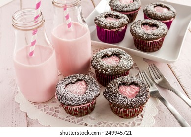 Chocolate cupcakes with heart shaped cutout on top filled with pink frosting and two bottles of strawberry milk sitting on white lace doily with two forks - Powered by Shutterstock