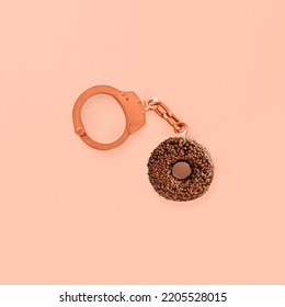 Chocolate Donut As A Part Of Handcuffs On Isolated Pastel Coral-pink Background. Abstract Concept Of Sugar Addiction Or Craving For Sweets. Eating Unhealthy Carbs Food. Minimal Flat Lay.