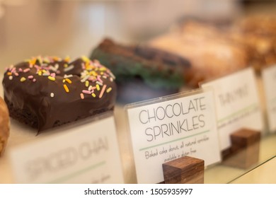 Chocolate Donut With Sprinkles In Bakery Display Case With Other Tempting Doughnuts, Focus On Chocolate Doughnut  And Description Tag Label, 