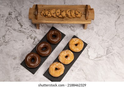 Chocolate Doughnuts And White Sugar Glaze On Two Slate Stone Plates, And Wooden Tray With Cookies Overhead View And Marble Table.