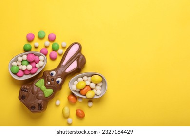 Chocolate Easter bunny, halves of egg and candies on yellow background, flat lay. Space for text