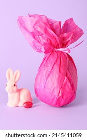 Chocolate Easter Egg Wrapped In Paper And Bunny On Lilac Background