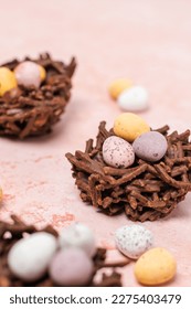 Chocolate Easter nests with mini eggs and hazelnuts. Nests are on a light pink background with mini eggs scattered around the image. 