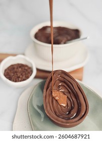 A chocolate egg shell being filled with creamy chocolate, with a bowl of chocolate ganache and cocoa nibs in the background on a wooden board.