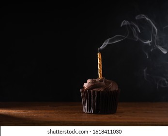 Chocolate Flavour Cupcake With Single Birthday Candle With Flame Blown Out With Flowing Smoke Set On Wooden Table,  And Dark Backdrop