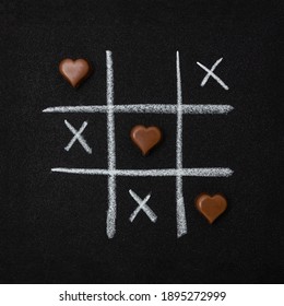 Chocolate Hearts And Grid Of Tic Tac Toe On Black Grainy Cardboard. Love Game Concept. Square Orientation.