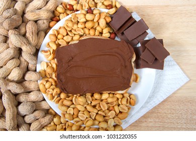 Chocolate Peanutbutter Spread On Bread, Open Face Sandwich On Plate With Pieces Of Chocolate, Shelled Peanuts And Whole In Shell Peanuts On Wood Table. Tasty Combination Of Chocolate And Peanuts