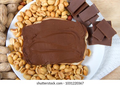 Chocolate Peanutbutter Spread On Bread, Open Face Sandwich On Plate With Pieces Of Chocolate, Shelled Peanuts And Whole In Shell Peanuts On Wood Table. Tasty Combination Of Chocolate And Peanuts