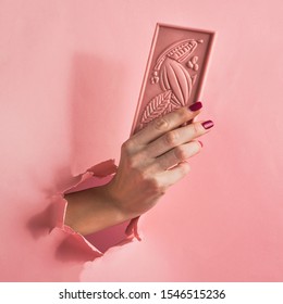 Chocolate Pink Bar Holds A Female Hand On A Pink Background Burst Paper Hole. Food Concept Art. Square Frame