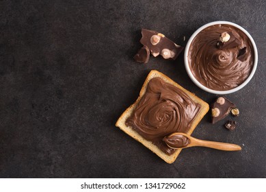 Chocolate spread on slice of bread with spoon, melted cream white bowl and pieces of chocolate with nuts on a stone table. Popular desser food. Top view.