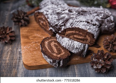 Chocolate yule log Christmas cake is a traditional French Christmas dessert with Mocha cream. Surrounded by fir-tree branches, cones, and berries, and covered in powdered sugar - Shutterstock ID 775518286