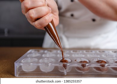 Chocolatier pours chocolate into molds. Chef in white apron using pastry bag filling hot melt chocolate into silicone mold. Concept for making homemade chocolate dessert in modern clean kitchen.