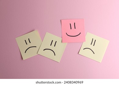 Choice concept. Sticky note with happy emoticon among beige papers with sad emojis on pink background, top view