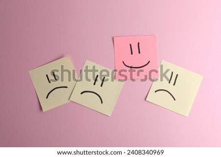 Choice concept. Sticky note with happy emoticon among beige papers with sad emojis on pink background, top view