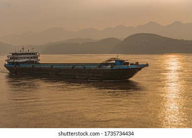 Chongqing, China - May 8, 2010: Evening Light On Yangtze River. Black-Blue Barge Sails On While Golden Sunset Reflected By Brown Water In Front Of Shoreline With Forested Hills.