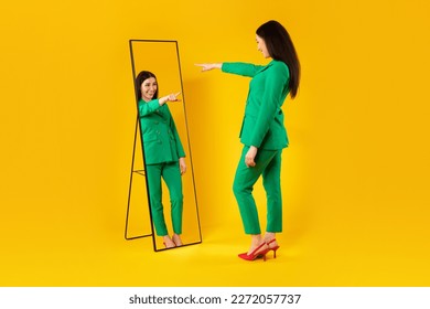 I choose myself. Excited young stylish lady pointing finger at her reflection in mirror, enjoying her new outfit, posing on yellow background, full length, free space. Self confidence concept - Shutterstock ID 2272057737