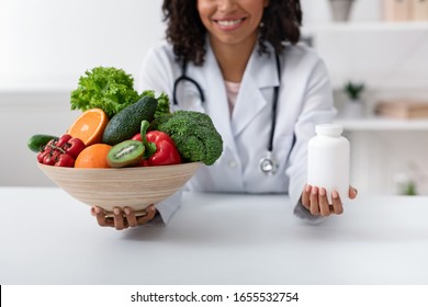 Choose quality healthy food. Afro woman nutritionist holding bowl with fresh fruits and vegetables and bottle of pills, close up, clinic interior