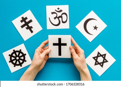 Choose Religion Concept. Hand With Catholic Cross Near World Religions Symbols On Blue Background Top View