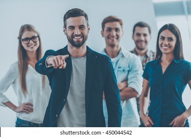 I Choose You For My Team! Handsome Young Man Pointing You And Smiling While Group Of Happy Young People Standing On Background 