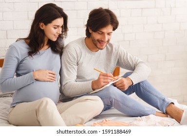 Choosing Baby Name. Young Pregnant Couple Naming Their Future Child, Writing Options To Notebook While Sitting On Bed At Home