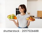 Choosing between healthy or unhealthy food. Young woman holding plates with fruits and sweets, looking at healthy snack with smile, deciding what to eat, standing in kitchen interior
