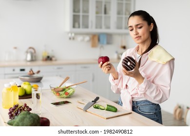 Choosing Between Healthy Or Unhealthy Food. Pensive Young Woman Holding Red Apple And Chocolate Donut, Looking At Sweet Snack With Suspicion, Deciding What To Eat, Standing In Kitchen, Free Space