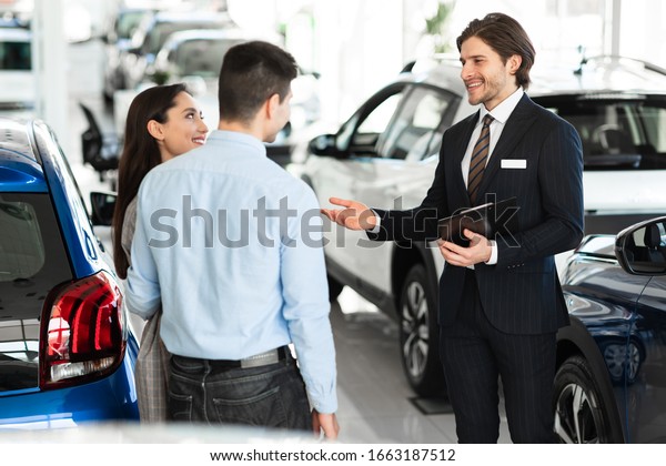 Choosing Car. Salesman\
Showing And Selling Auto To Couple Customers Standing In Luxury\
Automobile Showroom