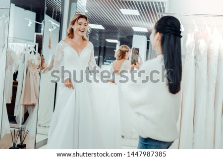 Choosing dresses. Beautiful smiling bride and her supportive bridesmaid taking pictures of a wedding dress in the shop.