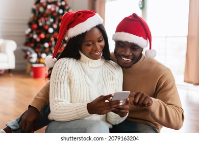 Choosing Festive Present. Smiling African American lady in Santa Claus hat showing her smartphone to boyfriend, guy pointing at screen, sharing gadget in decorated living room. Discount And Sale - Powered by Shutterstock