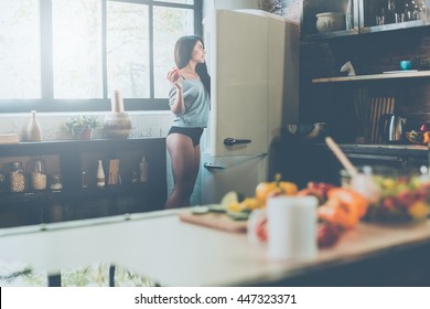 Choosing the freshest veggies for salad. Beautiful young African woman looking into the fridge and holding red pepper while standing in kitchen at home