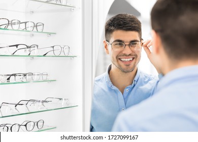 Choosing Glasses Concept. Portrait of handsome bearded guy picking new specs at optical shop, looking at mirror
