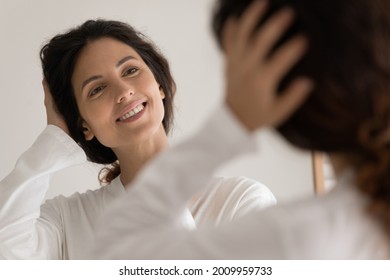 Choosing image for today. Reflection of happy smiling young hispanic woman in bathrobe touching head. Attractive millennial female caress long curly hair satisfied with pretty healthy look at mirror