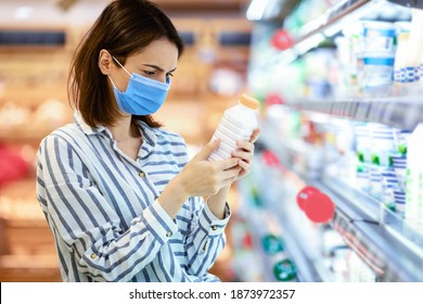 Choosing Milk. Female Customer In Medical Surgical Mask Holding Bottle Of Milk Or Yoghurt From The Fridge, Looking At Dairy Products, Standing Near Refrigerator Aisle, Checking Expiry Date