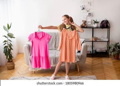 Choosing New Outfit. Cheerful Female Teenage Holding Two Nice Dresses At Modern Apartment