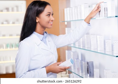 Choosing The Right Medicine For You. Beautiful Young African Woman In Lab Coat Choosing Medicine While Standing Near The Self In Drugstore 