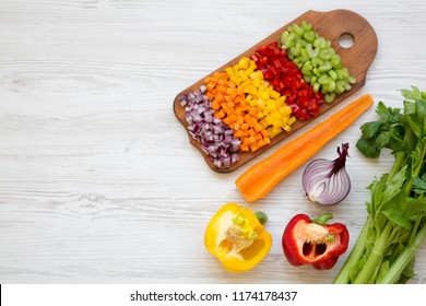 Chopped fresh vegetables (carrot, celery, red onion, red and yellow peppers) arranged on cutting board on white wooden table, overhead view. Flat lay, from above, top view. Copy space.