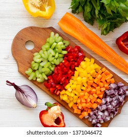 Chopped Fresh Vegetables (carrot, Celery, Onion, Colored Peppers) Arranged On Cutting Board On A White Wooden Surface, Top View. Overhead, From Above, Flat Lay.