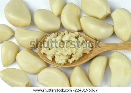 Chopped garlic in wooden spoon surrounded by peeled garlic cloves ready for cooking on a white background