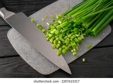 Chopped Pieces Of Green Onion Stalks And Sharp Metal Knife On Gray Plastic Board. Slices Of Fresh Green Scallion On Dark Gray Wooden Table. Cutting Shallots Or Chive, Seasonal Greenery. Close Up.