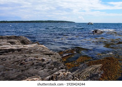 Choppy Ocean Waters Of Maine, Lobster Fishing Boat In The Background, Rocks In The Foreground. Blue Cloudy Sky