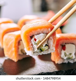 Chopstick holding Sushi Rolls Set with Salmon and Cream Cheese and Cuccumber on Black Slate Plate Close Up. Uramaki, Nori Maki or Futomaki Sushi with Trout Fillets, Soy Sauce and Wasabi - Powered by Shutterstock