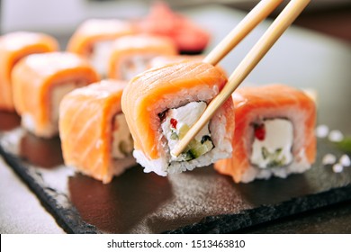 Chopstick holding Sushi Rolls Set with Salmon and Cream Cheese and Cuccumber on Black Slate Plate Close Up. Uramaki, Nori Maki or Futomaki Sushi with Trout Fillets, Soy Sauce and Wasabi - Powered by Shutterstock