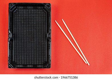 Chopsticks And Empty Rectangular Black Plastic Container For Products On A Red Background. Plastic Container For Sushi Set Delivery. Take Away Lunch Box. Top View. Flat Lay. Copy Space