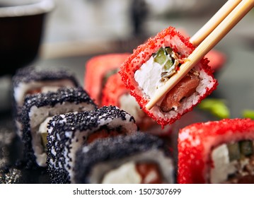 Chopsticks holding maki sushi roll with rice, cream cheese, salmon, eel, cucumber, flying fish caviar. Nori maki rolls with raw trout, red and black tobiko on natural dark stone background close up - Powered by Shutterstock