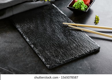 Chopsticks On Empty Black Sushi Plate. Ginger And Wasabi Sauce In Bowl On Dark Table. Asian Cuisine Idea. Traditional Condiment For Japanese Food. Oriental Restaurant Table Setting